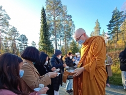 Bilder viser flere damer med munnbind på som står i rekke og gir mat til en munk som har på seg orange klær. Munken holder en bolle som damene legger mat i.