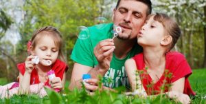 foto av en far og to barn som ligger i gresset og blåser såpebobler