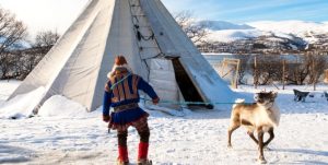 Bilde i farger. Et menneske med en fargerik drakt holder i et tau som er bundet rundt halsen på et reinsdyr. I bakgrunnen er det lavvo og snø.