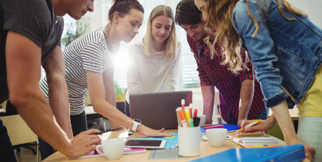 Fem voksne mennesker står rundt et bord. De har et møte.